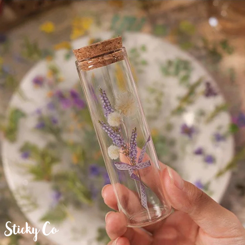 Glass tube with cork lid containing sticker flowers, held by a hand against a floral background. Brand 'Sticky Co' visible.