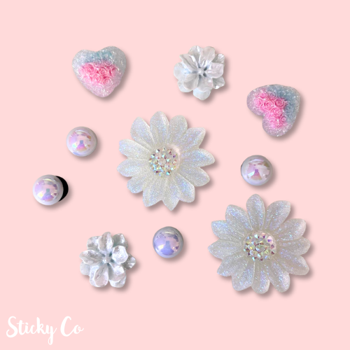 Decorative clog charms with floral and heart designs on a pink background, featuring the brand 'Sticky Co'.