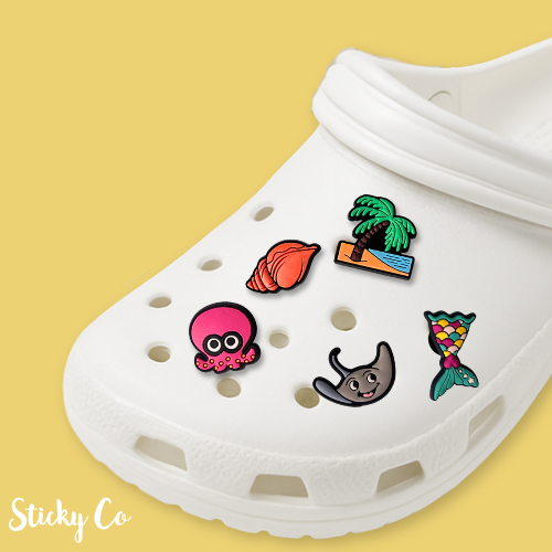 White clog with colorful charms on a yellow background, featuring the brand 'Sticky Co'.