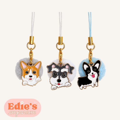 Three dog-shaped phone charms with pom-pom attachments on a white background, branded 'Edie's emporium'.